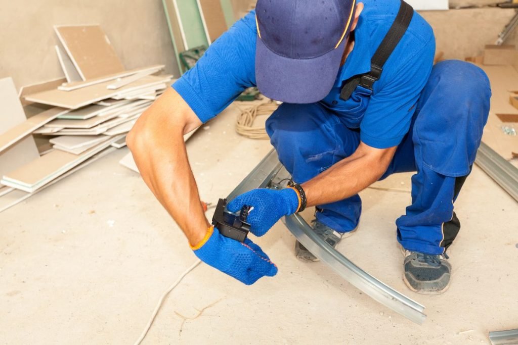 A construction worker in blue attire uses a tool to secure metal framing in a workshop filled with drywall materials.
