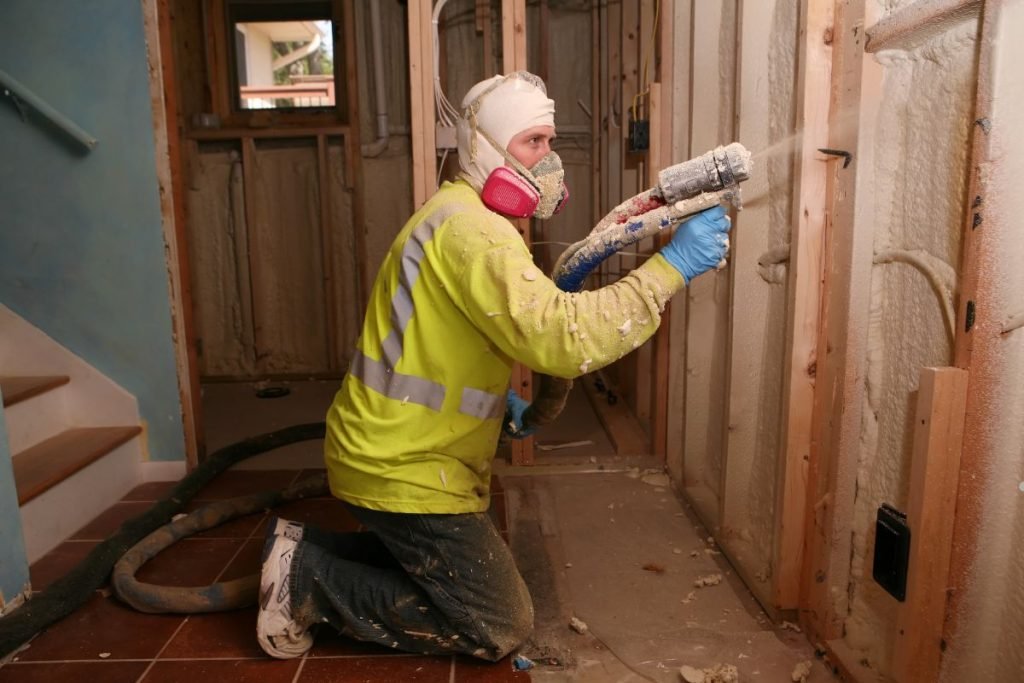 A worker in a bright yellow safety jacket sprays insulation material onto a wall in a partially constructed room.