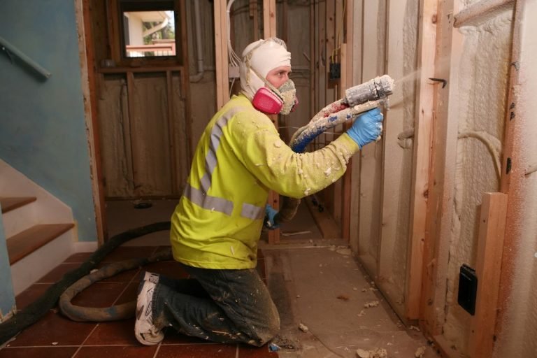 A worker in a bright yellow safety jacket sprays insulation material onto a wall in a partially constructed room.